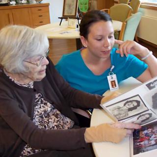 An elderly woman and a caregiver sitting at a table, looking through a photo album together.