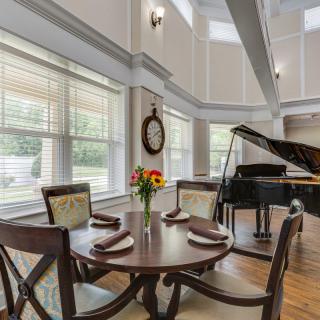 A spacious dining area featuring a round table set with napkins and flowers, adjacent to a grand piano and illuminated by natural light from large windows.