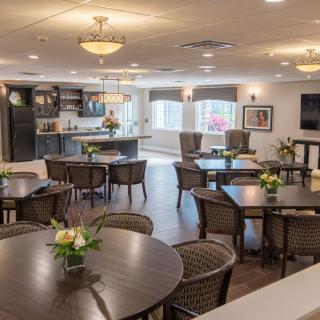 A well-lit dining area with multiple tables and chairs, featuring a kitchen in the background and decorative flowers on the tables.
