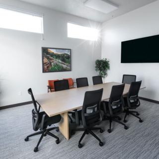 A modern conference room with a long table, several office chairs, a television on the wall, and decorative elements such as a potted plant and framed artwork.