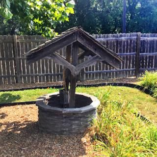 A rustic wooden well in a garden, surrounded by greenery and a wooden fence.