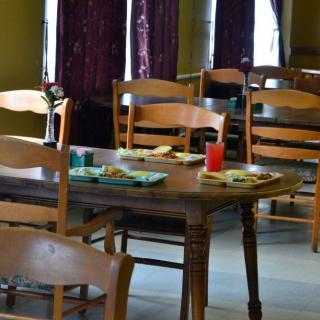 An empty dining room with wooden tables and chairs, set with plates and a red drink.