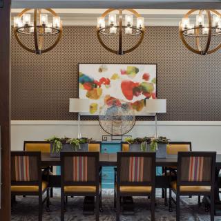A stylish dining room featuring a large table with chairs, under elegant chandeliers and colorful artwork on the wall.