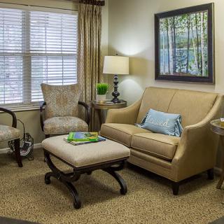 A cozy living room with neutral-toned furniture and decor, featuring a sofa, armchair, lamp, and artwork on the wall.
