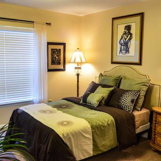 A cozy bedroom with a neatly made bed, decorated with green and brown bedding. The room features artwork on the walls, a lamp, and a window with blinds.