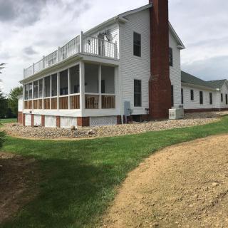 A white house with a brick chimney, featuring a large veranda and surrounded by a grassy lawn.