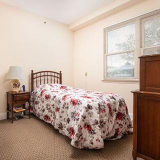 A cozy bedroom with a single bed covered in a floral bedspread, accompanied by a nightstand and lamp next to the window.