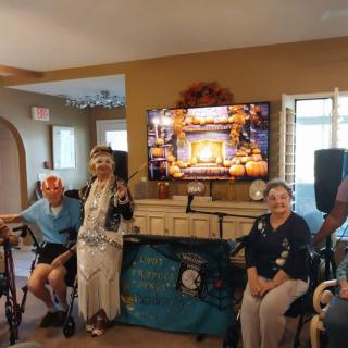 A group of elderly people gathered indoors, some in wheelchairs, smiling in front of a decorated TV.