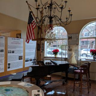 A cozy interior space featuring a piano, a chandelier, and an American flag, complemented by large windows and informational posters.