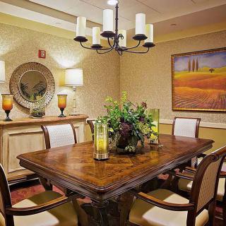 A cozy dining room with a wooden table and cushioned chairs, adorned with a chandelier and decorative elements like paintings, a mirror, and indoor plants.