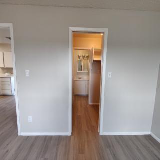 An empty room with a doorway leading to a hallway, featuring wooden flooring and white walls.