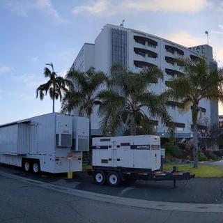 A large trailer and generator parked in front of a multi-story building surrounded by palm trees under a partly cloudy sky.