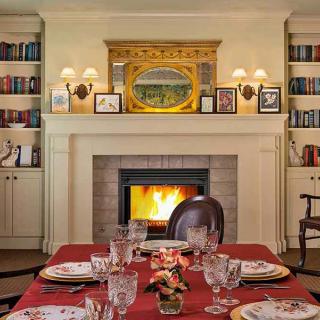A cozy dining room with a beautifully set table in front of a fireplace, surrounded by bookshelves.