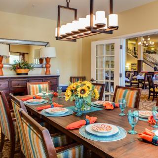 A vibrant dining room with a wooden table set with colorful plates and glasses, featuring a centerpiece of sunflowers. Striped chairs and a contemporary chandelier add to the cozy ambiance.