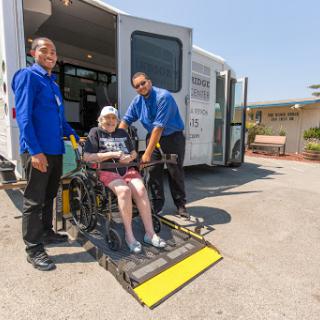 Caregivers assisting a senior in a wheelchair as they prepare for transportation.