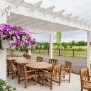 A charming outdoor patio with wooden tables and chairs under a white pergola, adorned with vibrant hanging flowers, overlooking a lush garden.