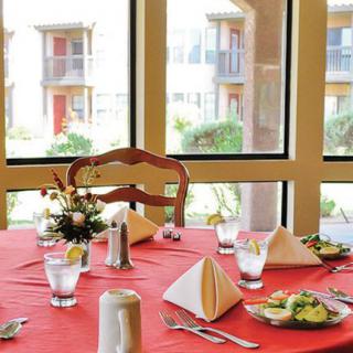 A dining table with a red tablecloth is elegantly set with folded napkins, plates, and glasses in a bright room with large windows.