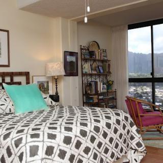A cozy bedroom with a patterned bedspread, bookshelf, and a view through large windows.