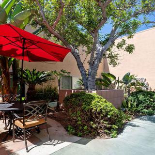 A cozy outdoor patio with a red umbrella, surrounded by lush greenery and a sunlit ambiance.