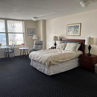 A cozy hotel bedroom featuring a queen bed, a small dining area, and large windows providing natural light.