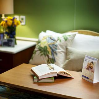 A cozy bedroom setup with an open book on a wooden table, surrounded by plush pillows and a bright bouquet of flowers under warm lighting.