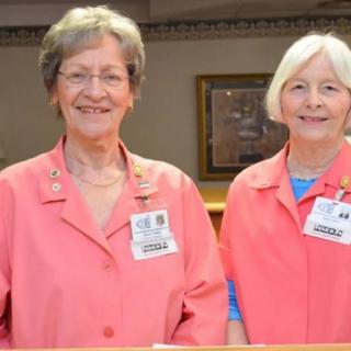 Two volunteers wearing pink uniforms stand at a hospital reception desk.