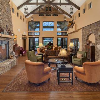 A spacious and rustic living room with a high ceiling, featuring wood beams, a stone fireplace, cozy seating, and large windows.