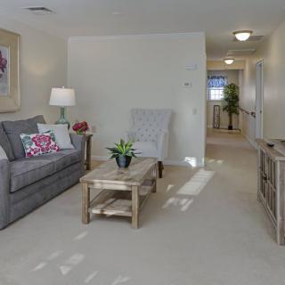 A cozy living room with a sofa, coffee table, and lamp, featuring light decor and carpeted flooring.