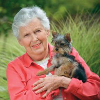 An elderly woman smiling and holding a small dog outdoors.