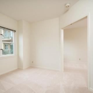 A bright, empty room with white walls and carpet, featuring a window and an open doorway.