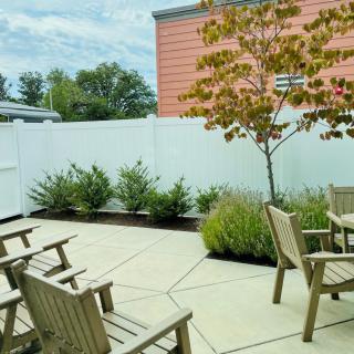 A peaceful patio area with wooden chairs, greenery, and a tree against a white fence.