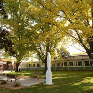 A peaceful garden area with a white statue, surrounded by trees and benches, adjacent to a building.