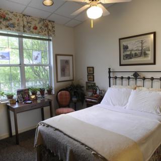 A cozy bedroom with a neatly made bed, a window letting in natural light, and decorative elements including framed pictures and a chair.