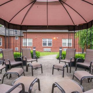 An inviting gazebo with comfortable seating surrounded by a garden and brick buildings.