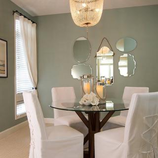 A modern dining room featuring a round glass table with white upholstered chairs, decorative mirrors on the wall, and soft ambient lighting.