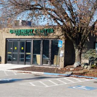A view of a business establishment featuring large glass windows and signage, surrounded by trees and a parking area.
