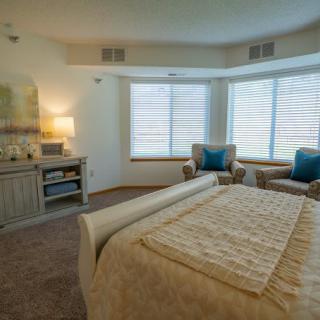 A cozy bedroom with a neatly arranged bed, armchairs by the window, and soft ambient lighting.