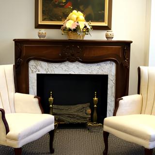 A cozy living room setting featuring a decorative fireplace flanked by two elegant chairs, with flowers and artwork above.