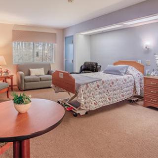 A well-furnished hospital patient room featuring a bed, seating area, and warm lighting.