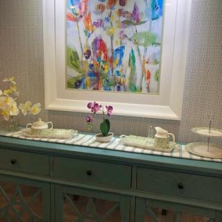 A decorated sideboard with teaware, flowers, and a colorful painting above.