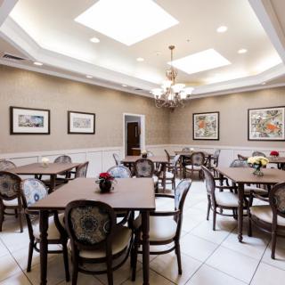 A well-lit dining room featuring multiple tables and chairs, decorated with framed artwork on the walls and a central chandelier under skylights.