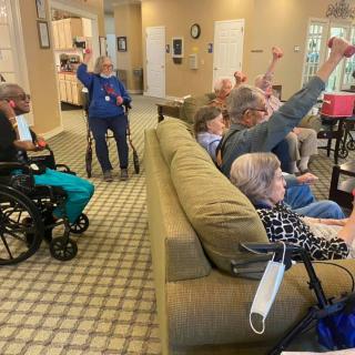 A group of seniors participating in an activity at a nursing home, with some sitting on a couch and others in wheelchairs, engaging enthusiastically.