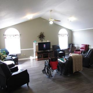 A cozy living room with various chairs, a television, large windows, and a wheelchair.