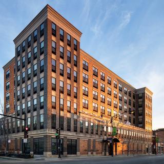A large multi-story brick building with numerous windows, set in an urban environment at sunset.