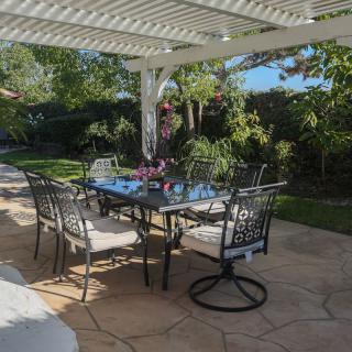 A sunny garden patio with a table and chairs under a pergola, surrounded by lush greenery.