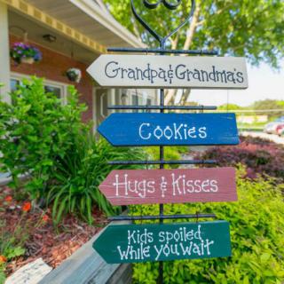 A colorful signpost in a garden setting directs visitors to 'Grandpa & Grandma's,' 'Cookies,' 'Hugs & Kisses,' and a 'Kids area while you wait.'