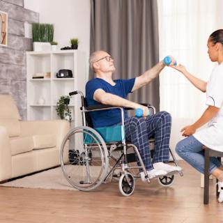 A caregiver assists an elderly person in a wheelchair with a rehabilitation exercise at home.