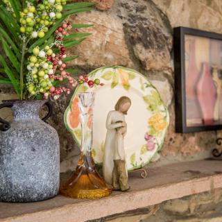 A decorative shelf display featuring a vase, a figurine, a colorful plate, and framed artwork against a stone wall.
