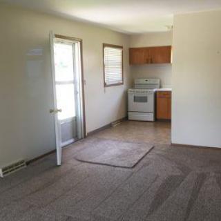 An empty apartment room with a small kitchen area featuring a stove and carpeted floor.