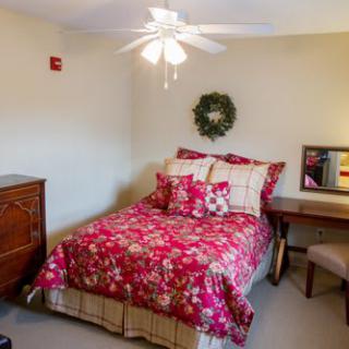 A cozy bedroom with floral red bedding and a ceiling fan, featuring a dresser, desk, and chair.